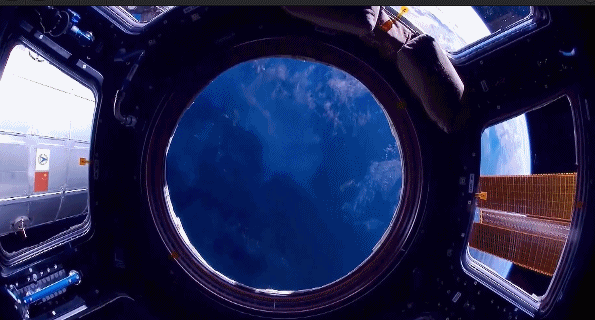 View of Earth through a round observation window from inside a space station, with solar panels visible outside.